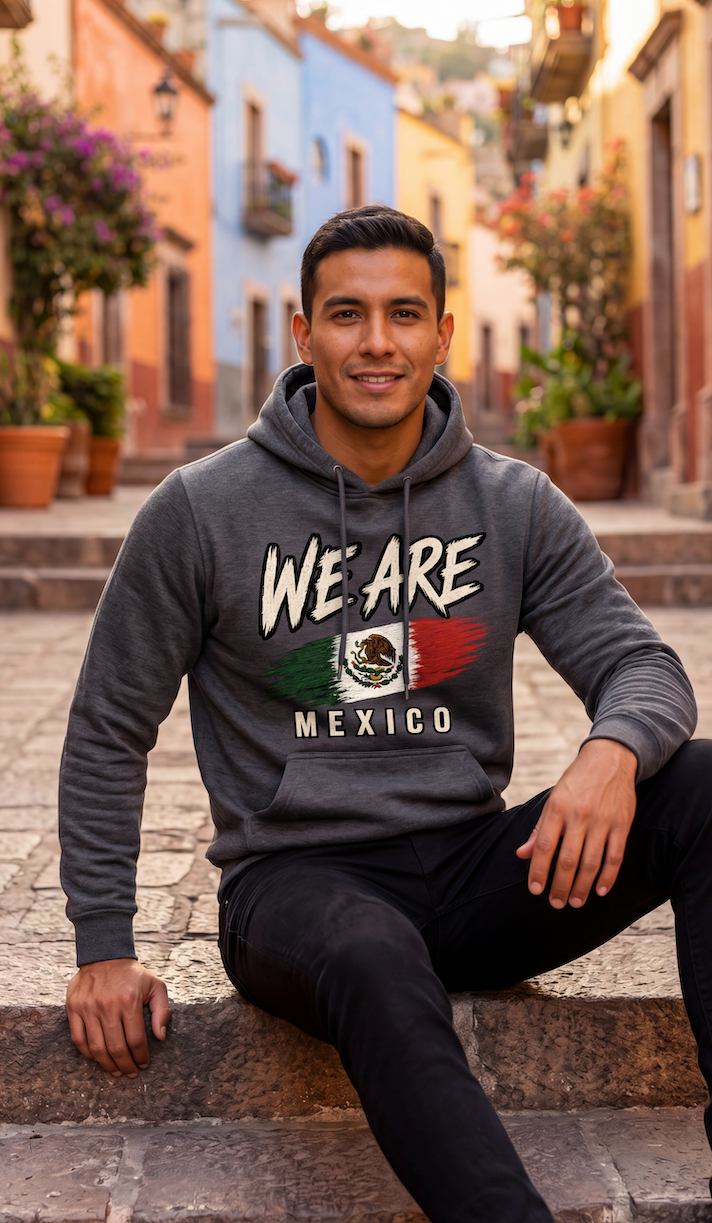A man sitting on stone steps in a cobblestone street wearing a dark gray hoodie with 'WE ARE MEXICO' and a Mexican flag graphic on the front, paired with black pants.