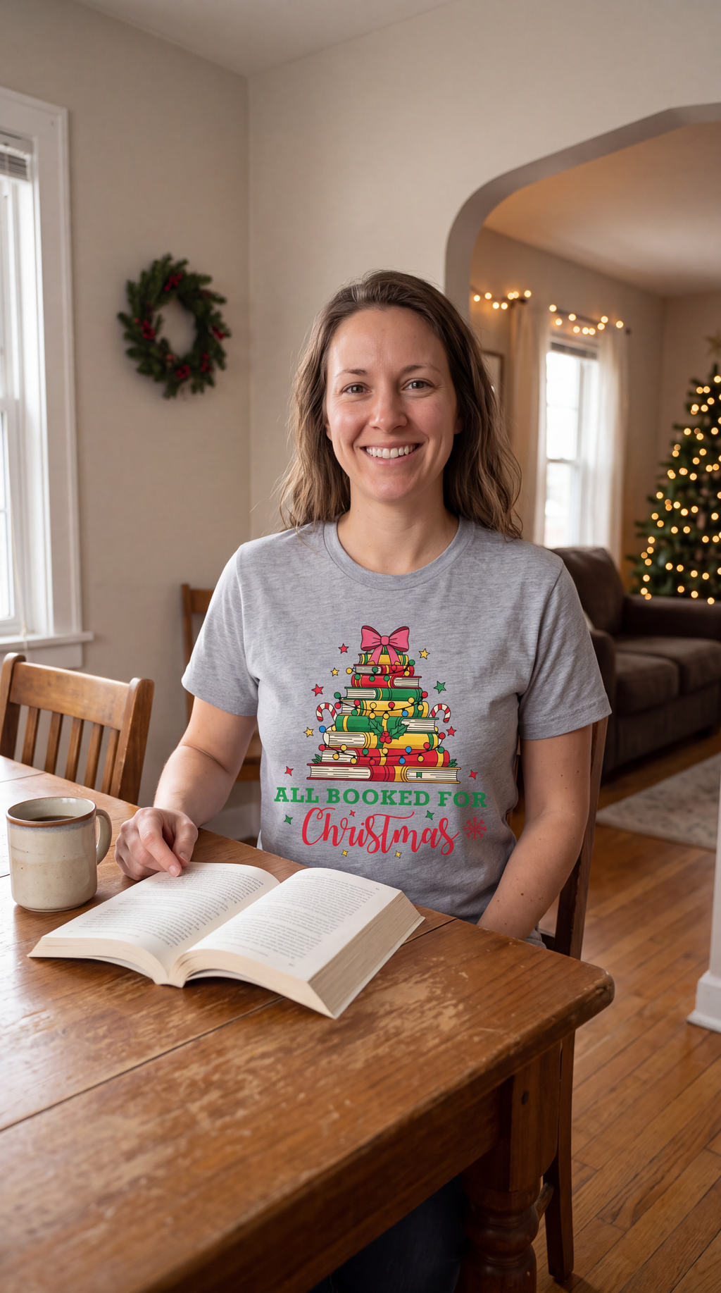 Woman wearing a heather gray All Booked for Christmas shirt while reading in a cozy holiday setting
