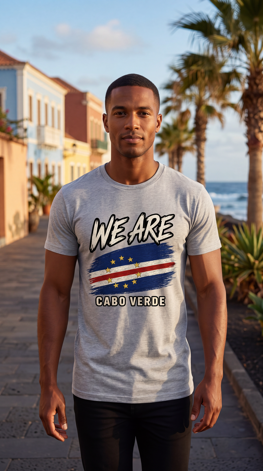 A man wearing a light gray heather t-shirt with a graphic of the Cabo Verde flag and the text 'WE ARE CABO VERDE' stands on a seaside walkway with palm trees and buildings in the background.