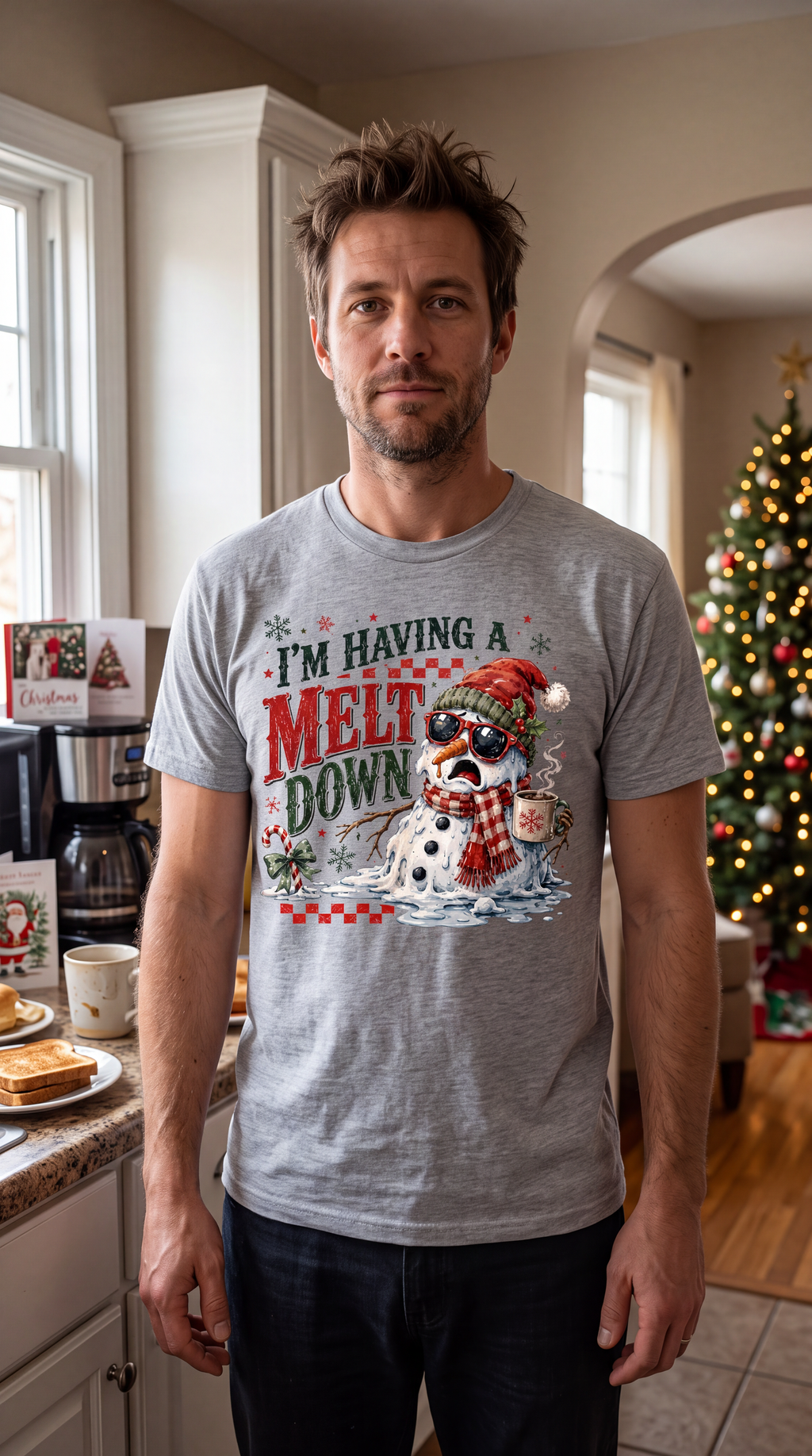 Man wearing heather gray I’m Having a Meltdown Christmas shirt indoors beside a decorated Christmas tree.