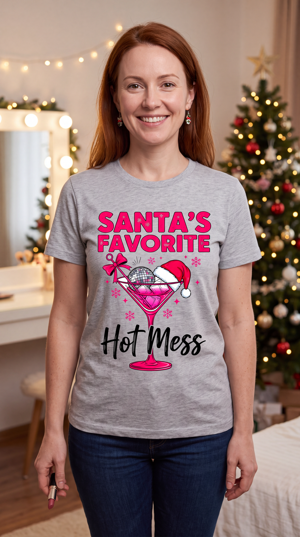 Woman wearing heather gray Santa’s Favorite Hot Mess Christmas shirt near a decorated Christmas tree.