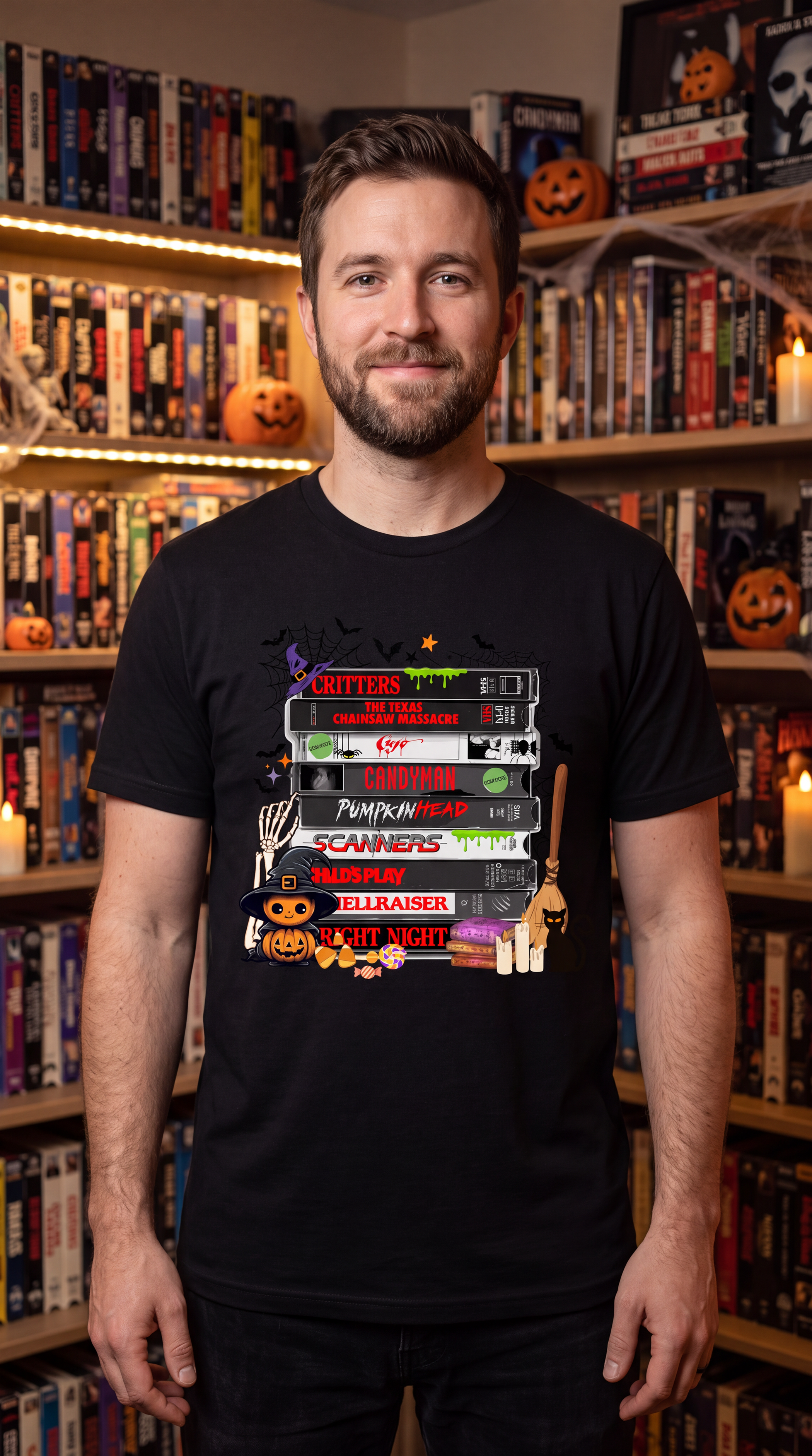 A man wearing a black graphic t-shirt featuring a stack of VHS tapes with pop culture movie titles, standing in front of bookshelves decorated for Halloween.