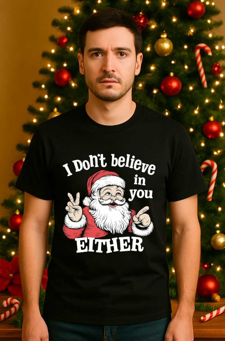 Man wearing a black Christmas t-shirt with a funny Santa graphic and the text ‘I Don’t Believe in You Either,’ standing in front of a decorated Christmas tree with ornaments and candy canes.