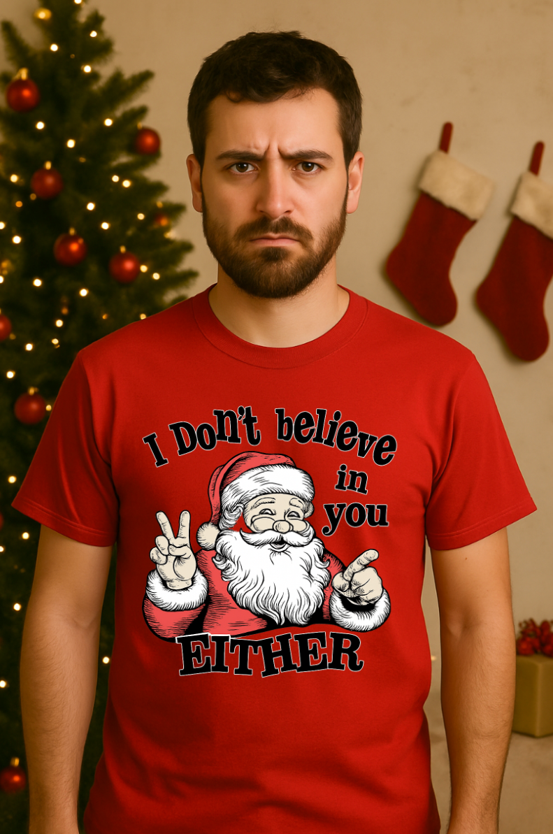 Man wearing a red Christmas t-shirt with a funny Santa graphic and the text ‘I Don’t Believe in You Either,’ standing in front of a decorated Christmas tree with stockings in the background.