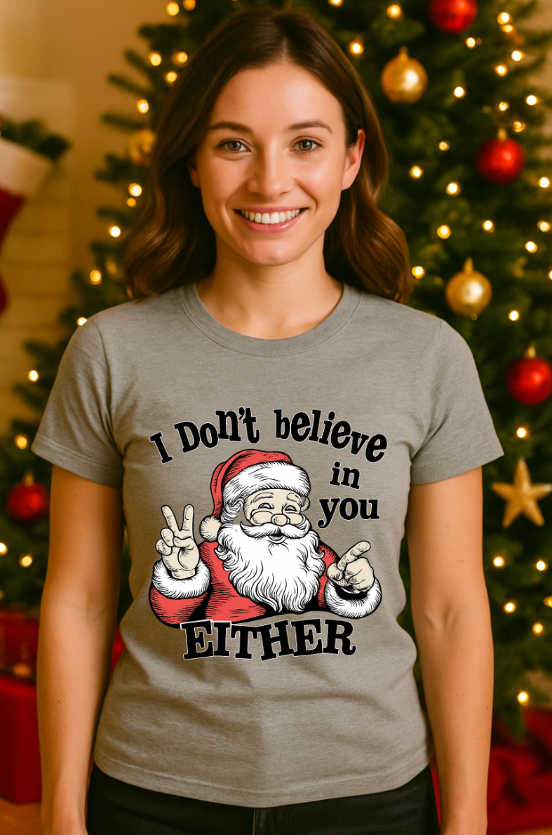 Woman wearing a gray Christmas t-shirt with a funny Santa graphic and the text ‘I Don’t Believe in You Either,’ standing in front of a decorated Christmas tree with lights and ornaments.