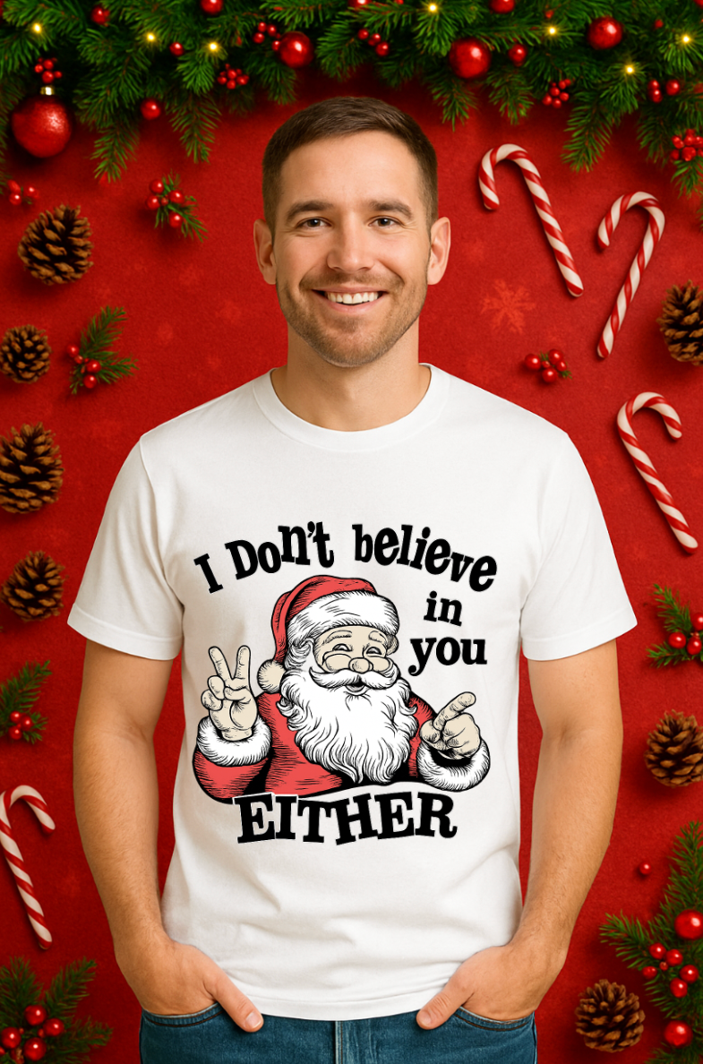 Man wearing a white Christmas t-shirt with a funny Santa graphic and the text ‘I Don’t Believe in You Either,’ standing in front of a festive red holiday background with candy canes, pinecones, and greenery.