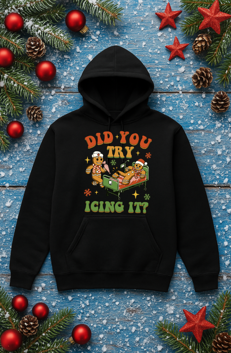 Black hoodie featuring a funny gingerbread “Did You Try Icing It?” Christmas graphic, displayed flat on a festive blue wooden background with ornaments, pine branches, and holiday decorations.