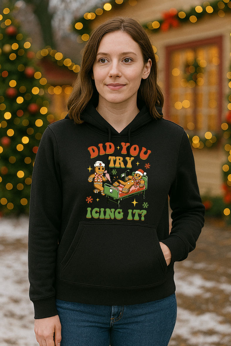 Woman wearing a black “Did You Try Icing It?” gingerbread Christmas hoodie, standing outside in front of holiday lights and festive decorations.