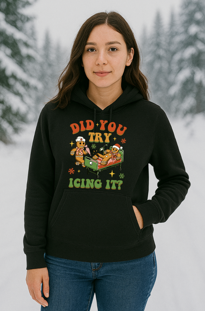 Woman wearing a black “Did You Try Icing It?” gingerbread Christmas hoodie, standing outside in a snowy winter setting with pine trees in the background.