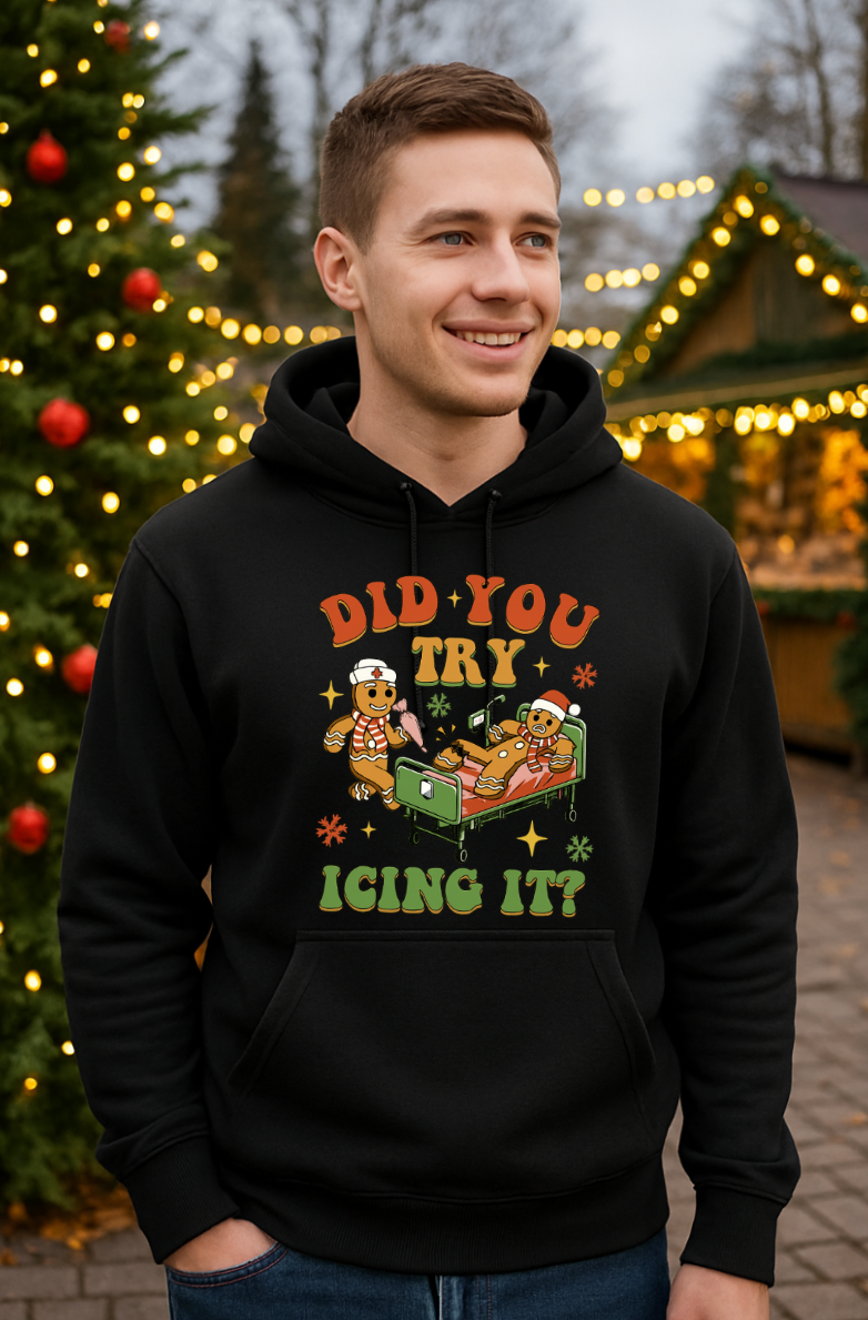 Man wearing a black “Did You Try Icing It?” gingerbread Christmas hoodie, standing outdoors in front of holiday lights and decorated trees.