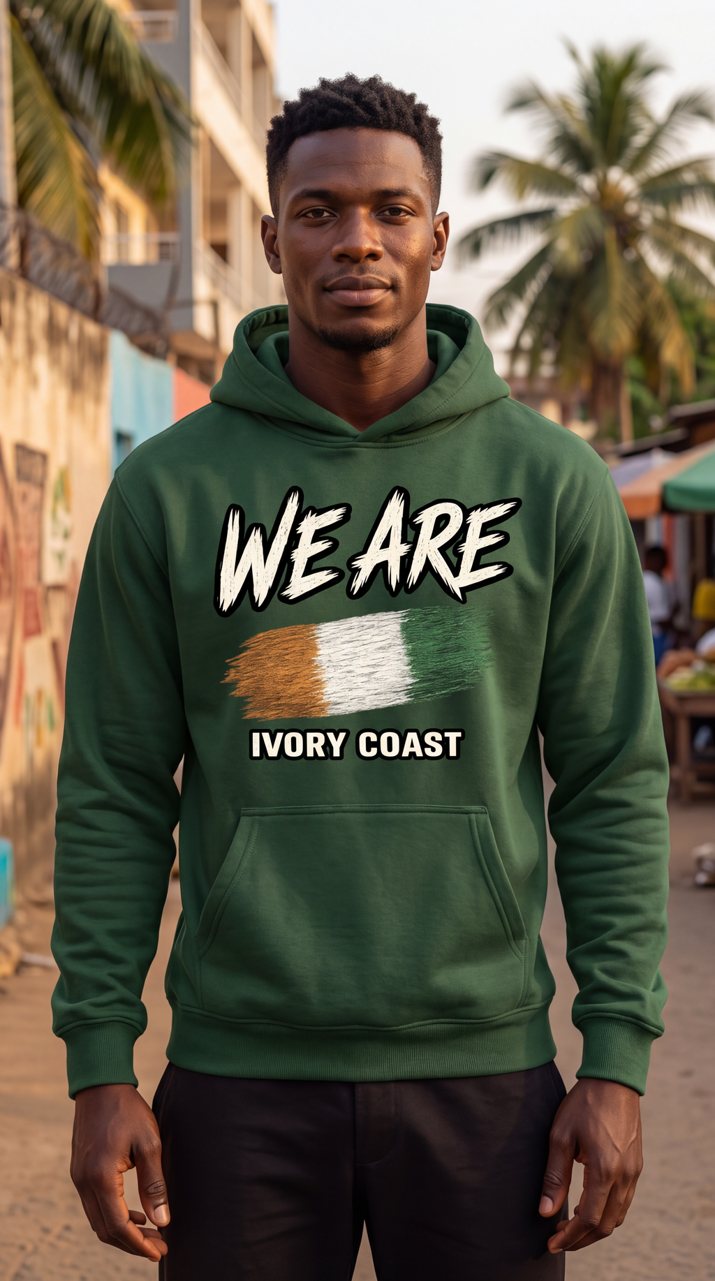 A man wearing a dark green hoodie with a graphic print that reads 'WE ARE' above the Ivory Coast flag and 'IVORY COAST' below, standing outdoors in a street setting with palm trees and buildings blurred in the background.
