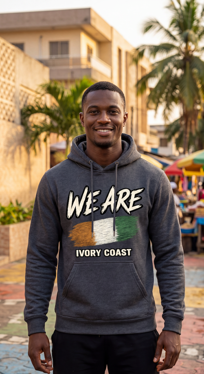 A smiling man wearing a dark gray heather hoodie with a graphic that reads 'WE ARE' above the Ivory Coast flag and 'IVORY COAST' below, standing outdoors in front of buildings and palm trees.