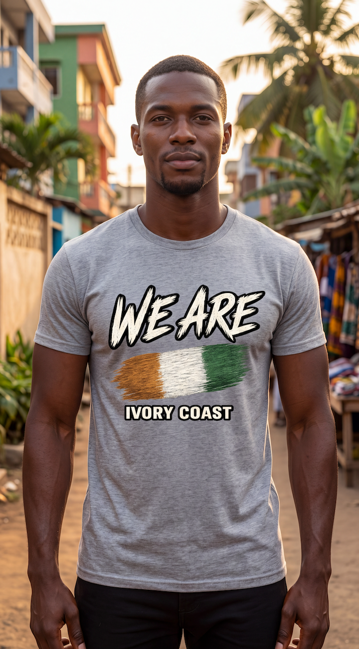A man wearing a heather gray t-shirt with a graphic that reads 'WE ARE' above the Ivory Coast flag and 'IVORY COAST' below, standing in an outdoor street setting.