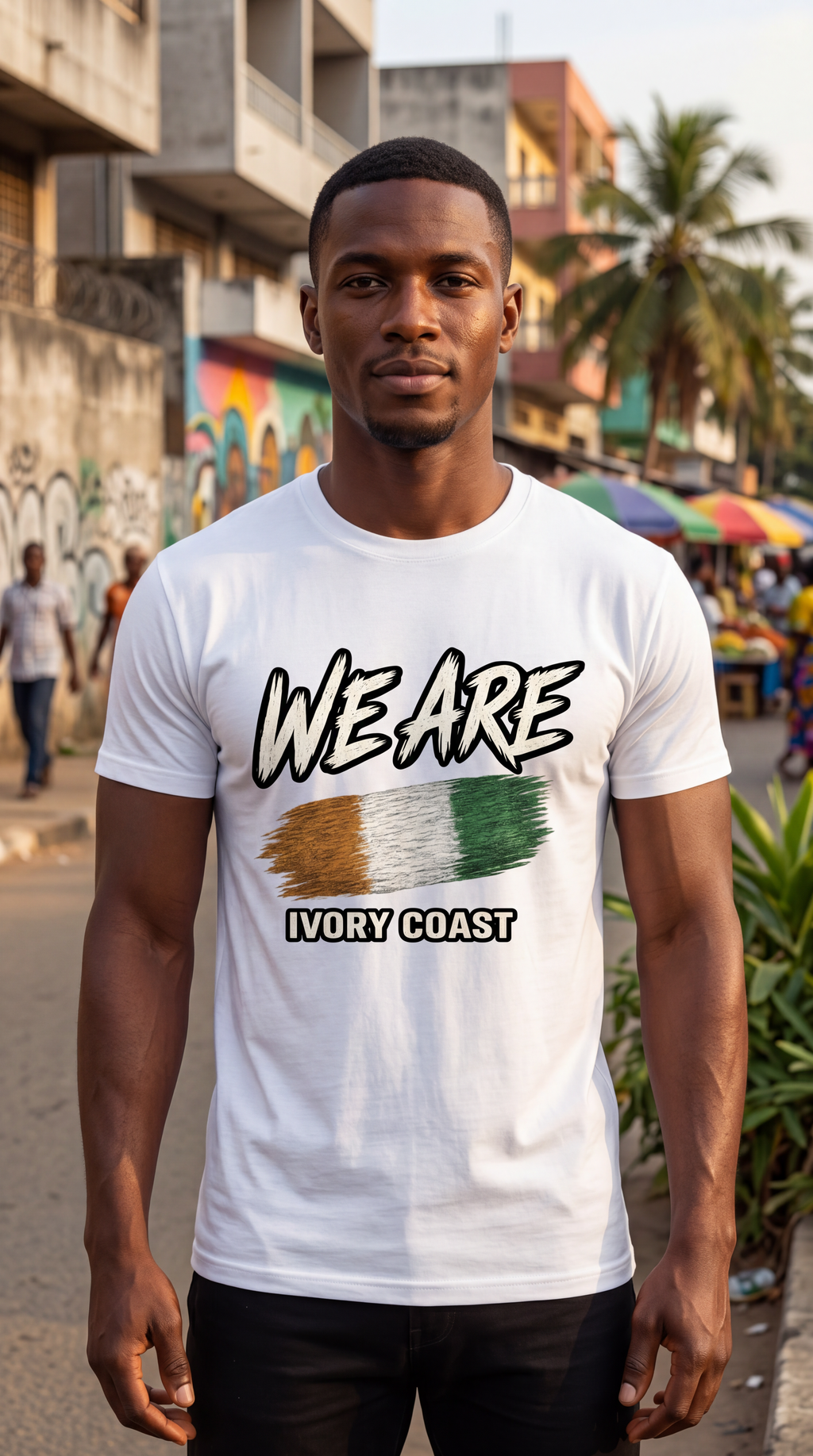 A man wearing a white graphic t-shirt with 'WE ARE IVORY COAST' and a stylized Ivorian flag design, standing in an outdoor street setting.