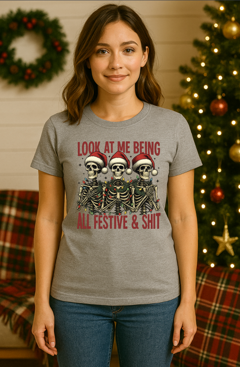 Woman wearing a gray Christmas skeleton t-shirt with three skeletons in Santa hats and the text “Look At Me Being All Festive & Shit,” standing in front of a decorated Christmas tree