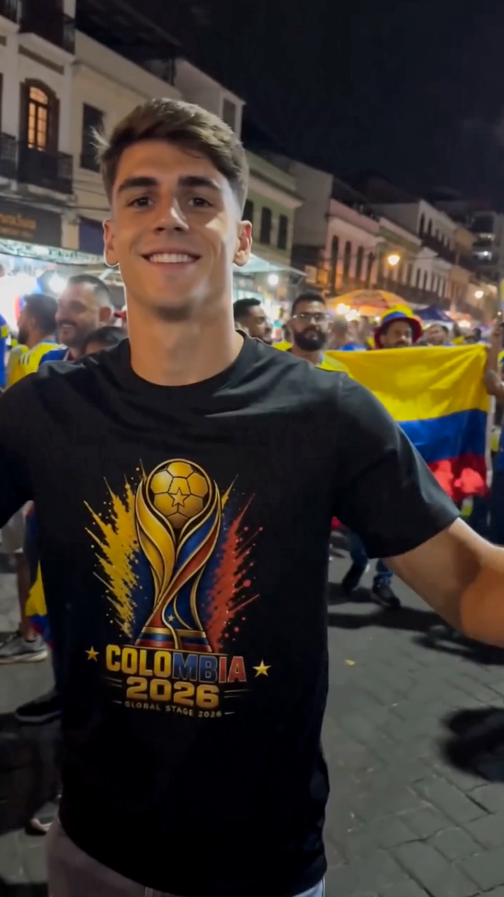 Man wearing black Colombia 2026 football fan shirt celebrating with Colombia supporters at night