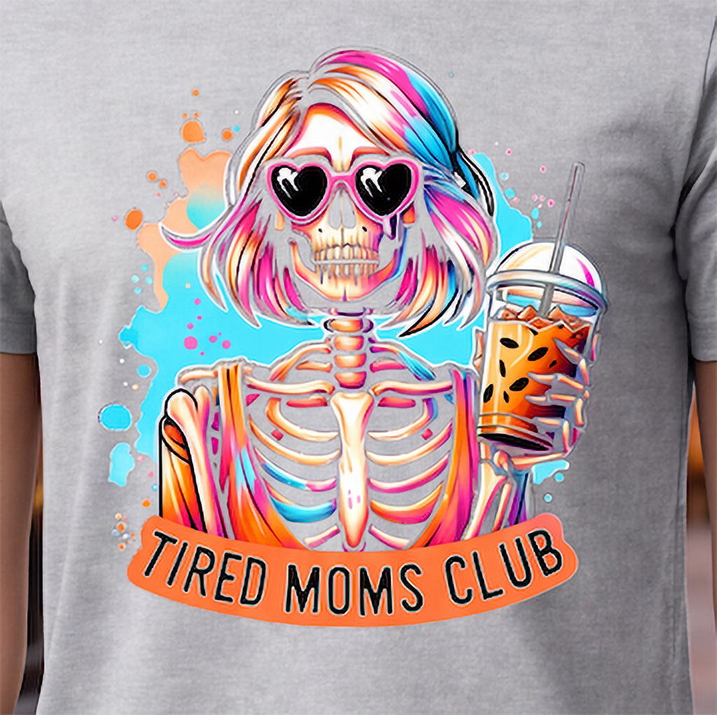 A close-up of a heather gray t-shirt featuring a colorful graphic of a skeleton with pink and blue hair, heart-shaped sunglasses, and a drink, with an orange banner reading 'TIRED MOMS CLUB'.