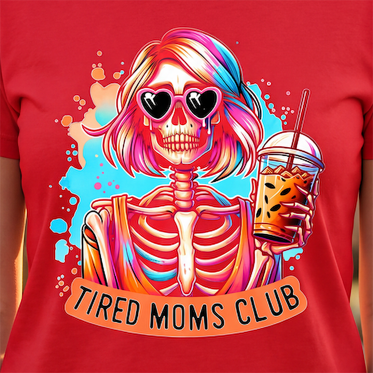 A red short-sleeve T-shirt shown on a model, featuring a colorful graphic of a cartoon skeleton with pink and blue hair, heart-shaped sunglasses, and an iced coffee, with the text 'TIRED MOMS CLUB' below.
