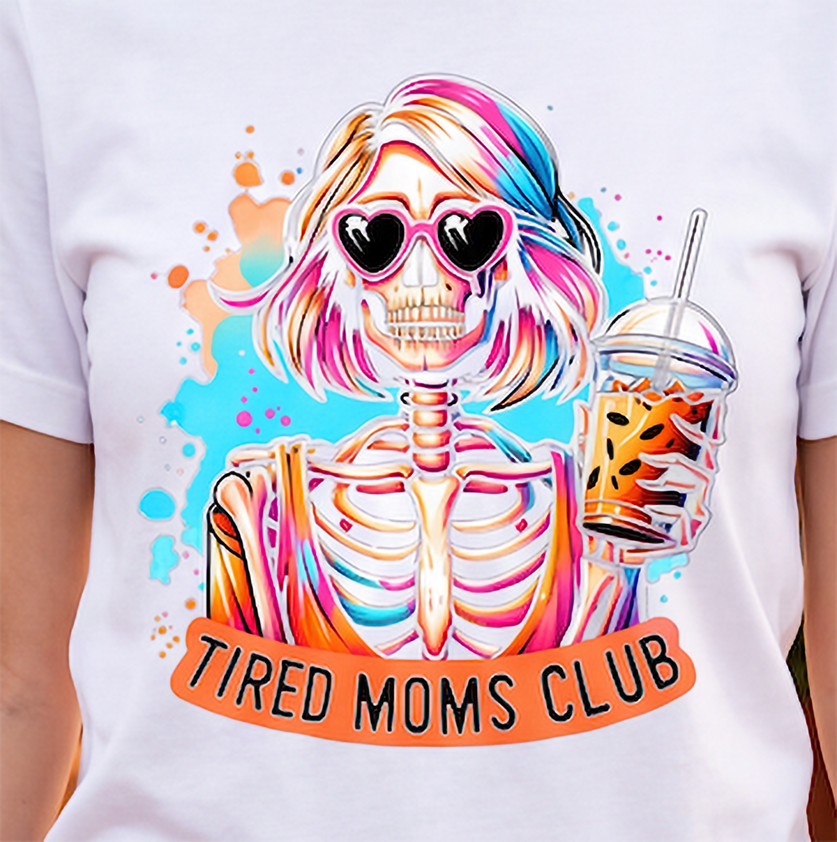 A white short-sleeve T-shirt featuring a colorful graphic of a cartoon skeleton woman with pink, blue, and orange hair, heart-shaped sunglasses, and an iced coffee, with an orange banner reading 'TIRED MOMS CLUB'.