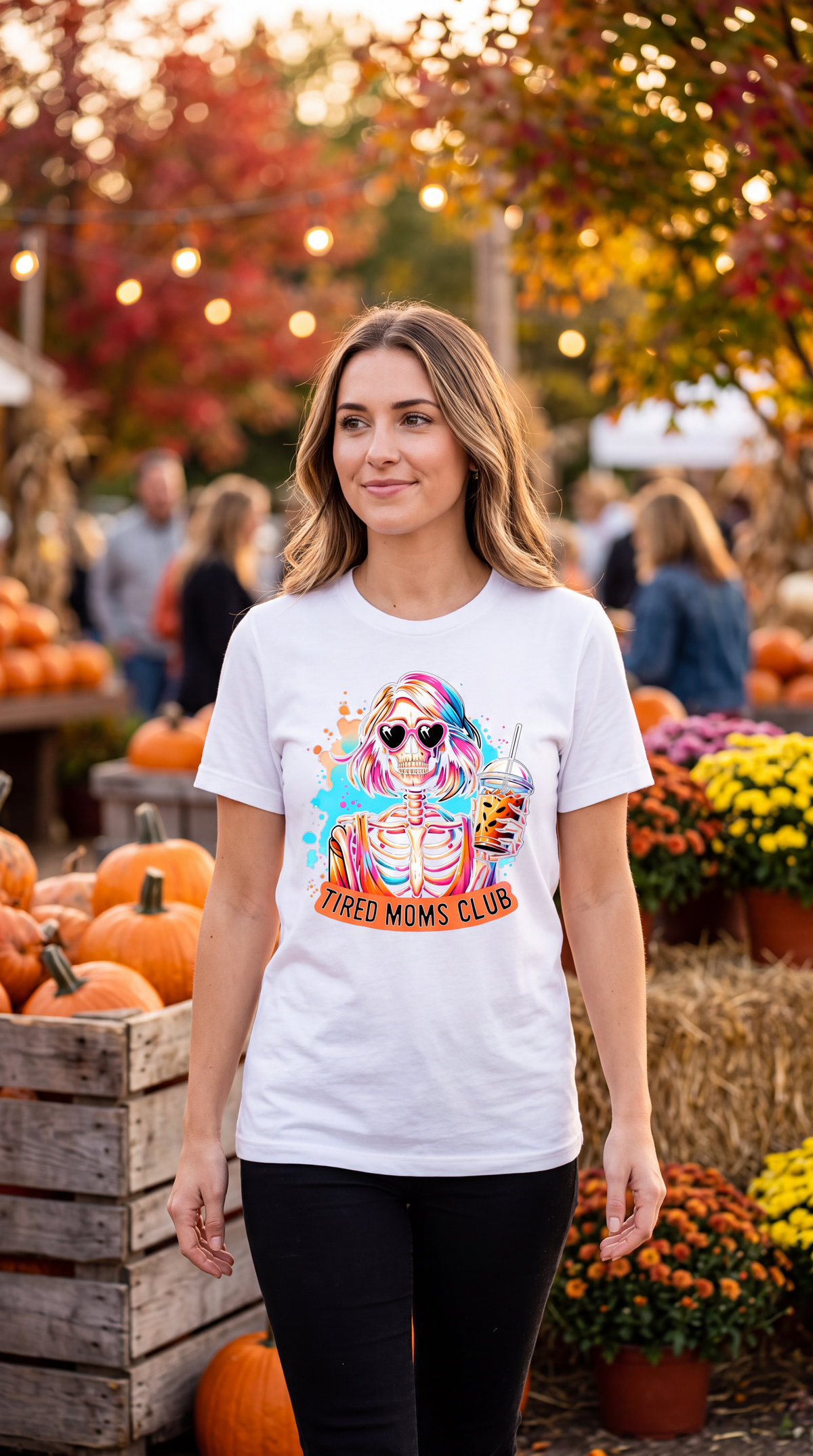 A woman wearing a white graphic t-shirt with a colorful 'Tired Moms Club' design, standing in an outdoor autumn pumpkin patch with pumpkins and fall foliage in the background.