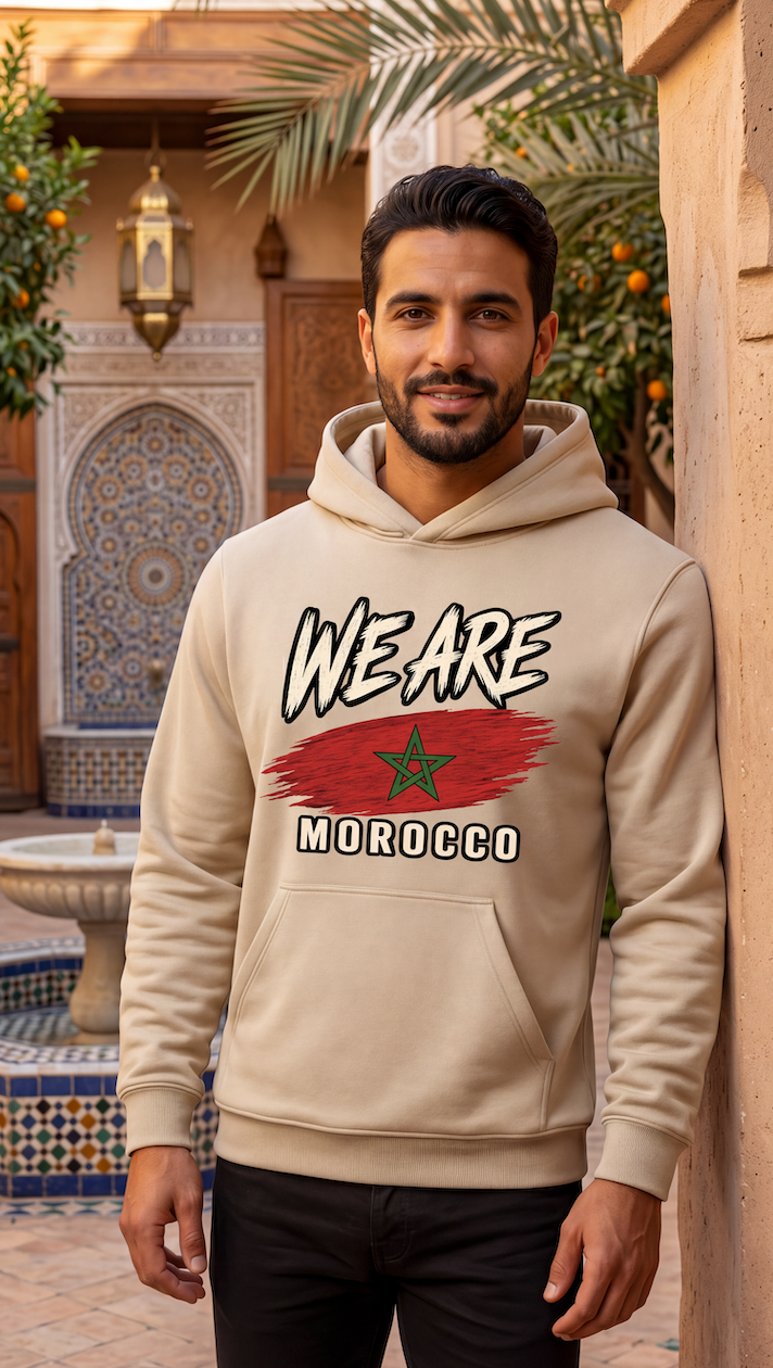 A man wearing a beige hoodie with a graphic print that reads 'WE ARE MOROCCO' and features a red brushstroke design with a green star, standing in a Moroccan courtyard.