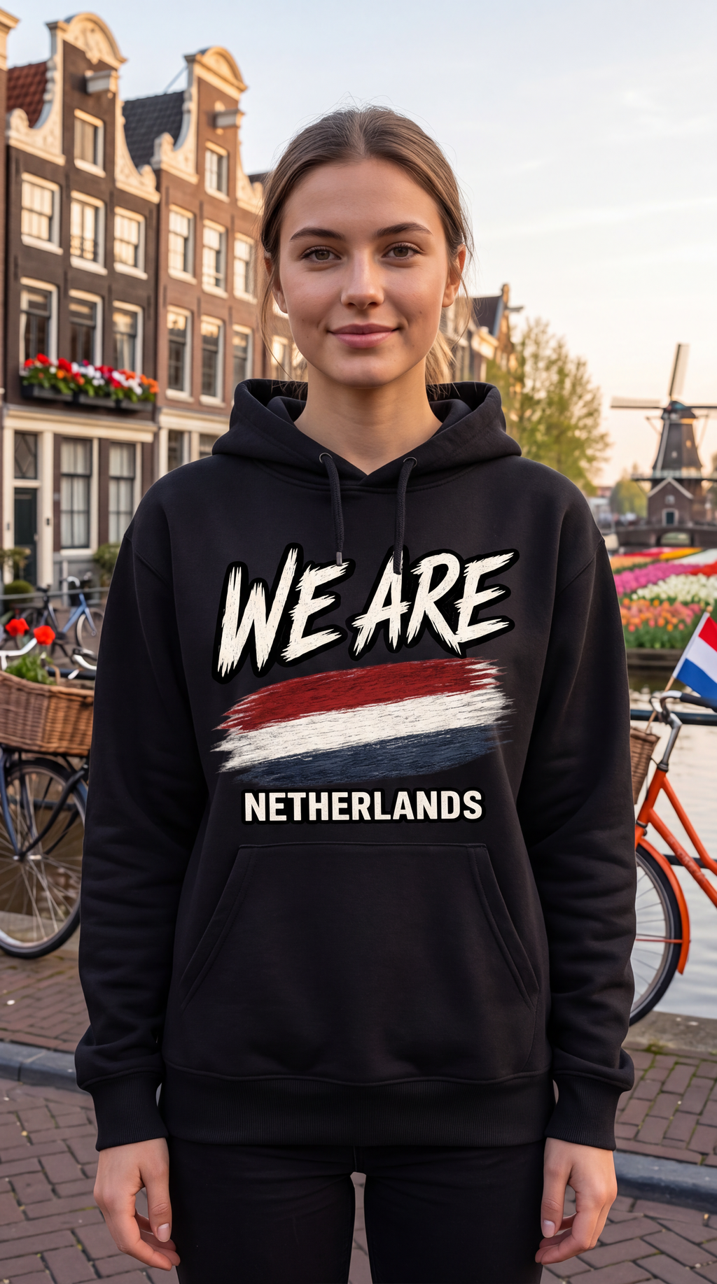 A woman wearing a black hoodie with 'WE ARE NETHERLANDS' and a red, white, and blue flag graphic, standing in front of a canal-side street with Dutch buildings and bicycles.