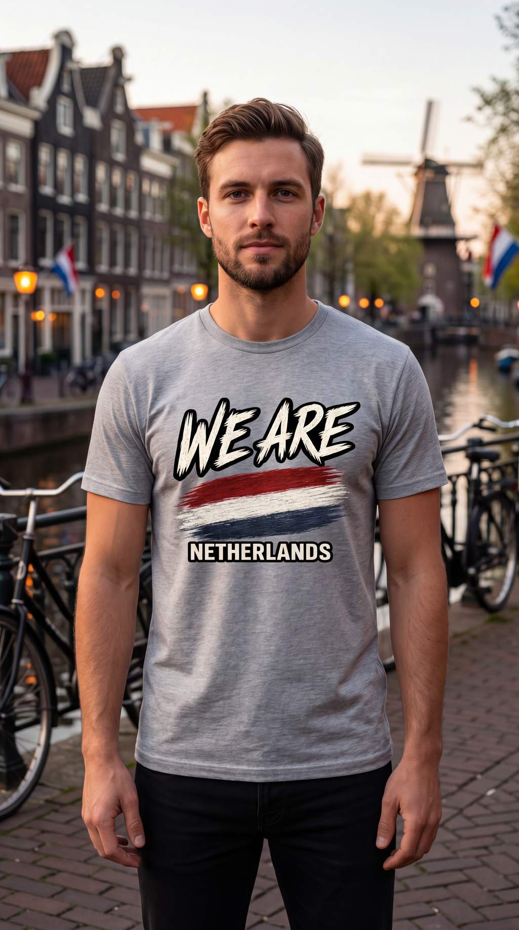 A man wearing a heather gray t-shirt with a 'WE ARE NETHERLANDS' graphic featuring the Dutch flag, standing in front of a canal with Amsterdam houses and a windmill in the background.