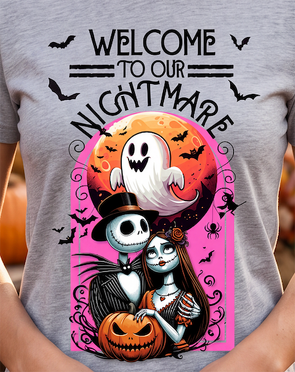 A person wearing a heather gray graphic T-shirt with a large Halloween design. The print reads “WELCOME TO OUR NIGHTMARE” above an illustrated ghost, a skeleton character in a top hat, a woman with long dark hair, a jack-o'-lantern, bats, and a pink moon background.