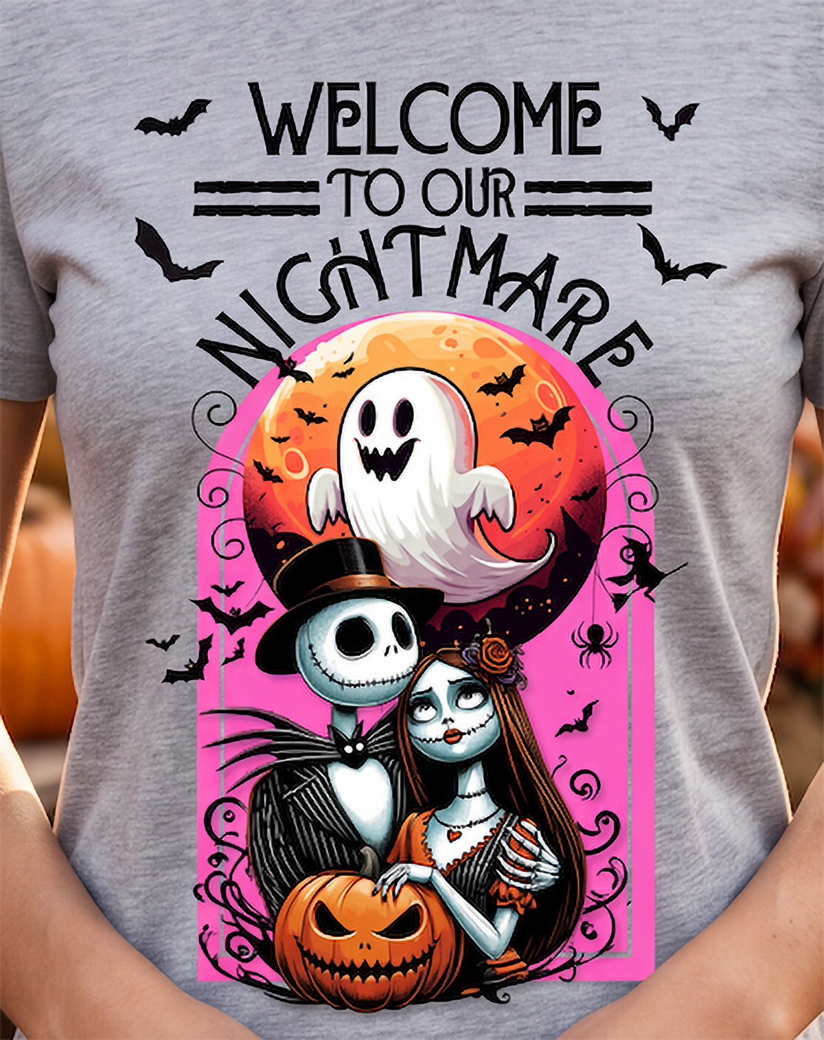 A person wearing a heather gray graphic T-shirt with a large Halloween design. The print reads “WELCOME TO OUR NIGHTMARE” above an illustrated ghost, a skeleton character in a top hat, a woman with long dark hair, a jack-o'-lantern, bats, and a pink moon background.