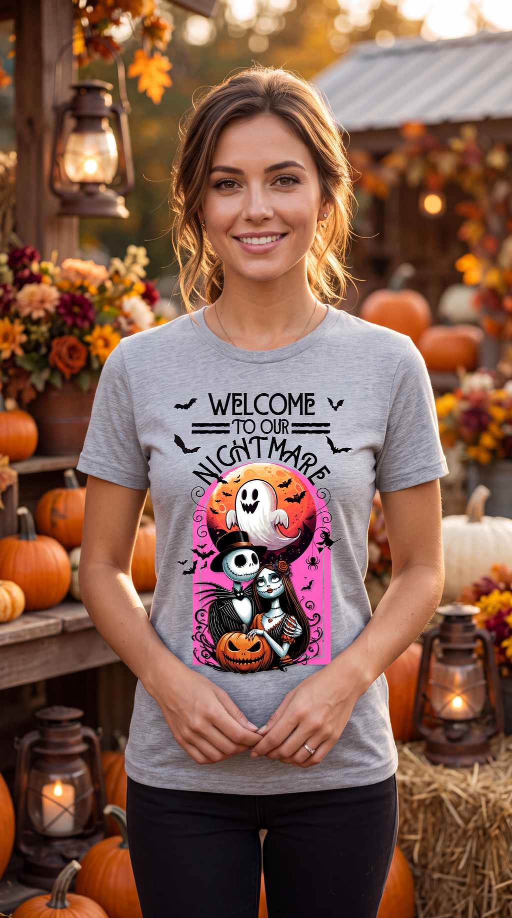 A woman wearing a heather gray graphic t-shirt with a Halloween-themed design that reads 'WELCOME TO OUR NIGHTMARE' above Jack Skellington, Sally, and a ghost. She is standing in a decorated autumn outdoor setting with pumpkins, lanterns, and fall foliage.