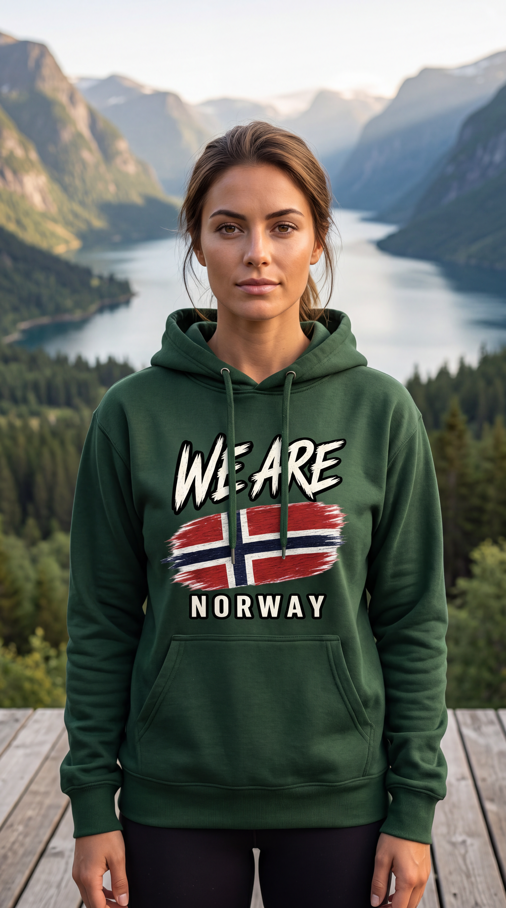 A woman stands outdoors in front of a mountain lake landscape wearing a dark green hoodie with a graphic that reads 'WE ARE NORWAY' and a Norwegian flag design on the chest.