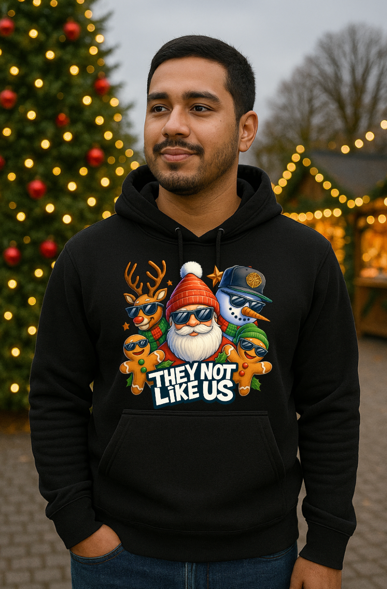 Man wearing a black Christmas hoodie with a funny holiday streetwear graphic featuring Santa, reindeer, snowman, and gingerbread characters, standing outdoors near a decorated Christmas tree with lights.