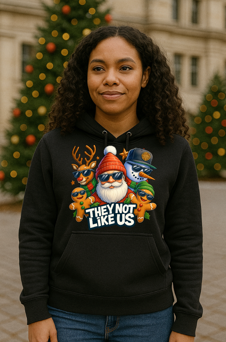 Woman wearing a black Christmas hoodie with a funny holiday streetwear graphic featuring Santa, reindeer, snowman, and gingerbread characters, standing outdoors in front of decorated Christmas trees.