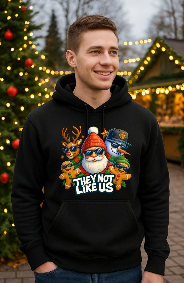 Man wearing a black Christmas hoodie with a funny holiday streetwear graphic featuring Santa, reindeer, snowman, and gingerbread characters, standing outdoors near a decorated Christmas tree with lights.
