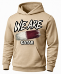 A beige pullover hoodie with a graphic print on the front. The design features the text 'WE ARE' in a white and black brush-stroke style above a stylized Qatar flag, and 'QATAR' in white block letters below.