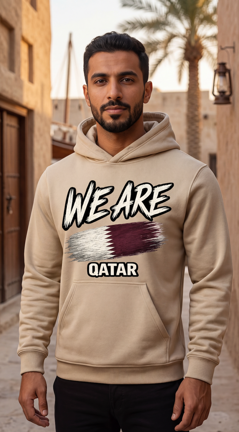 A man wearing a beige hoodie with a graphic print that reads 'WE ARE QATAR' and features the Qatar flag design, standing in an outdoor street setting.