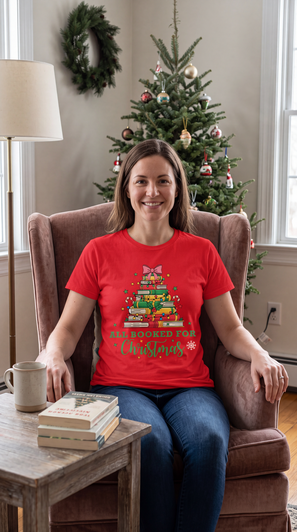 Woman wearing a red All Booked for Christmas book lover shirt in a cozy Christmas reading setting