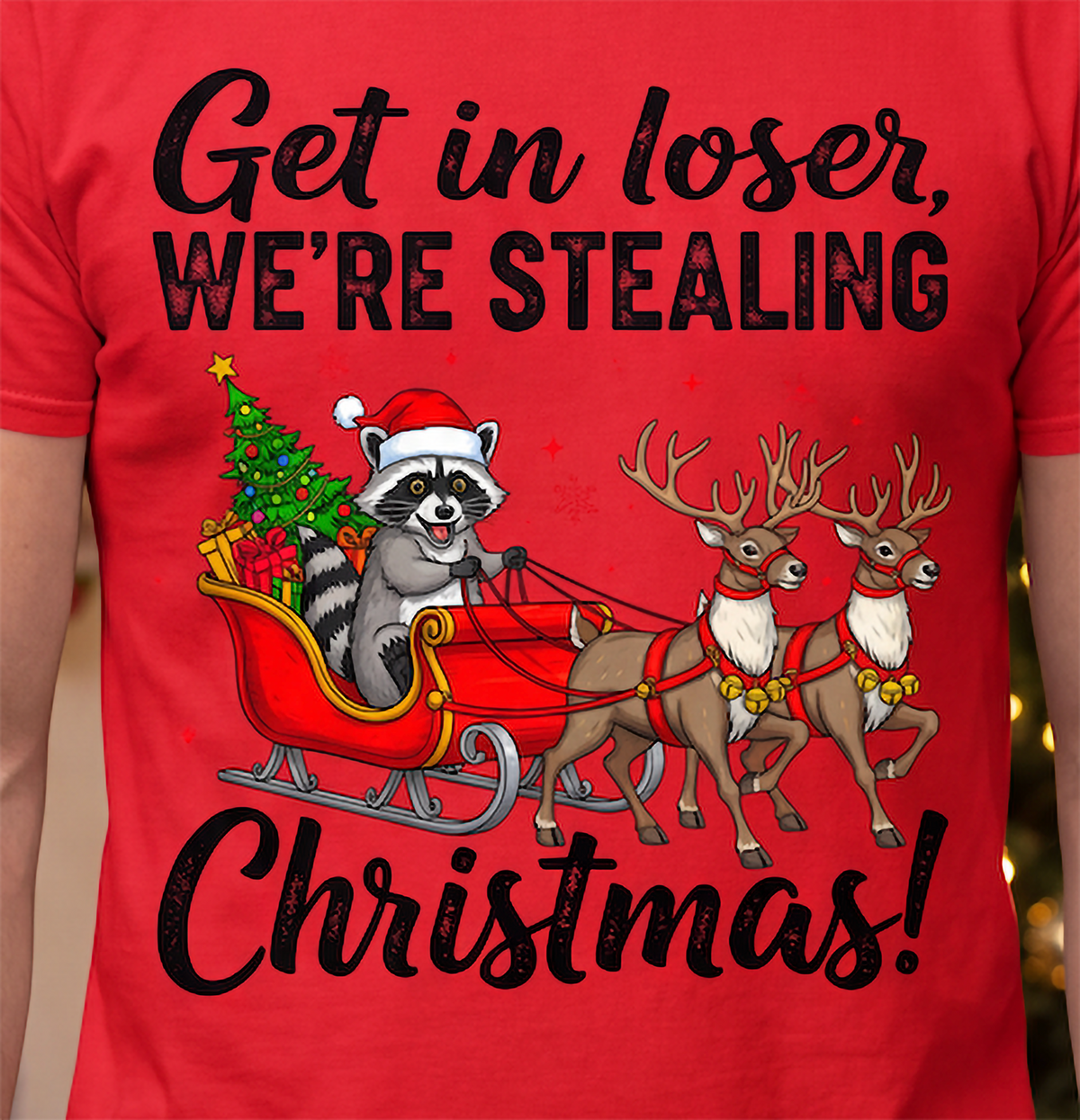 Close-up of Get in Loser We’re Stealing Christmas raccoon, sleigh and reindeer graphic on a red T-shirt.