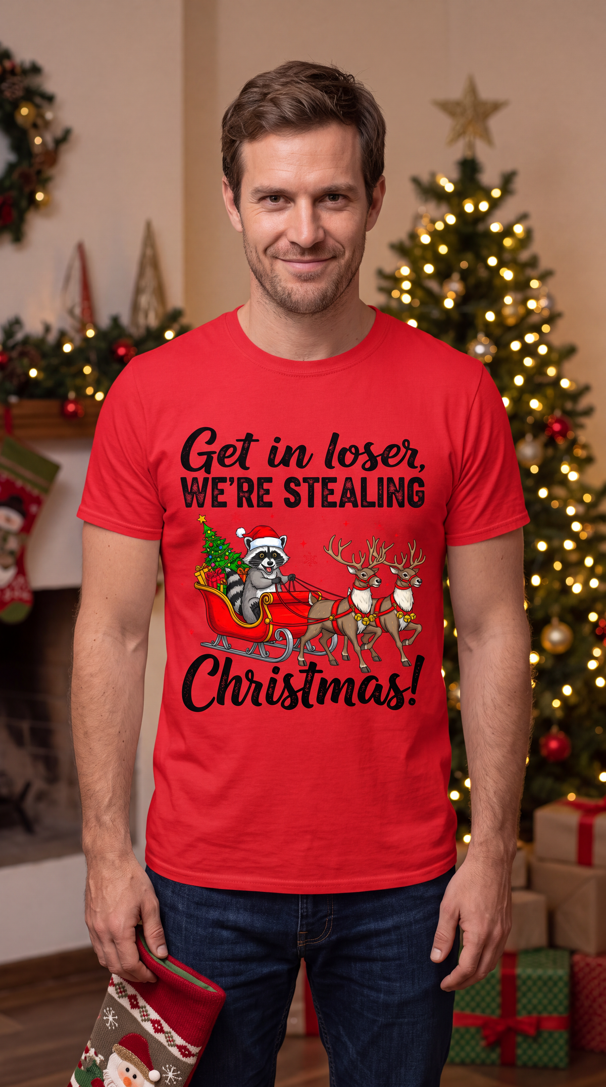 Man wearing a red Get in Loser We’re Stealing Christmas graphic tee beside Christmas decorations.
