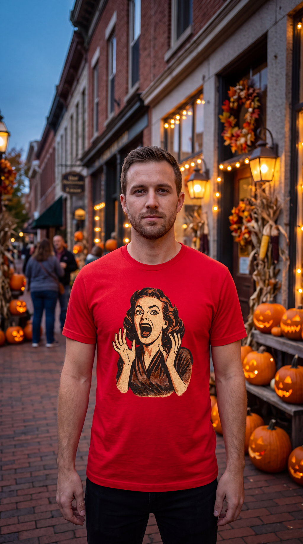 A man wearing a bright red graphic t-shirt stands on a brick street lined with Halloween decorations, including pumpkins and storefronts with warm lights.