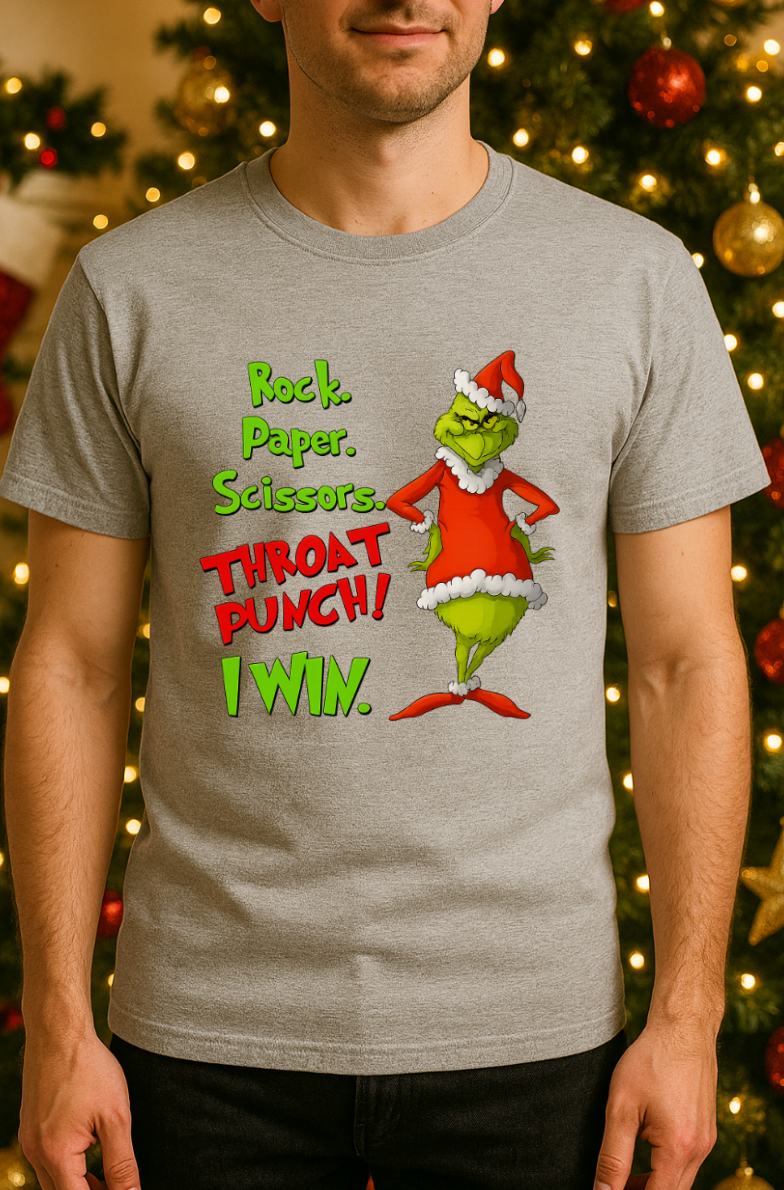 Man wearing a light gray Christmas graphic t-shirt featuring a green mischievous character in a red Santa outfit with the phrase “Rock. Paper. Scissors. Throat Punch! I Win.” standing in front of a decorated Christmas tree with warm holiday lights and ornaments.