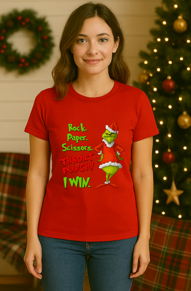 Woman wearing a red Christmas graphic t-shirt featuring a green mischievous character in a red Santa outfit with the phrase “Rock. Paper. Scissors. Throat Punch! I Win.” standing in a festive holiday setting with a decorated Christmas tree and wreath.