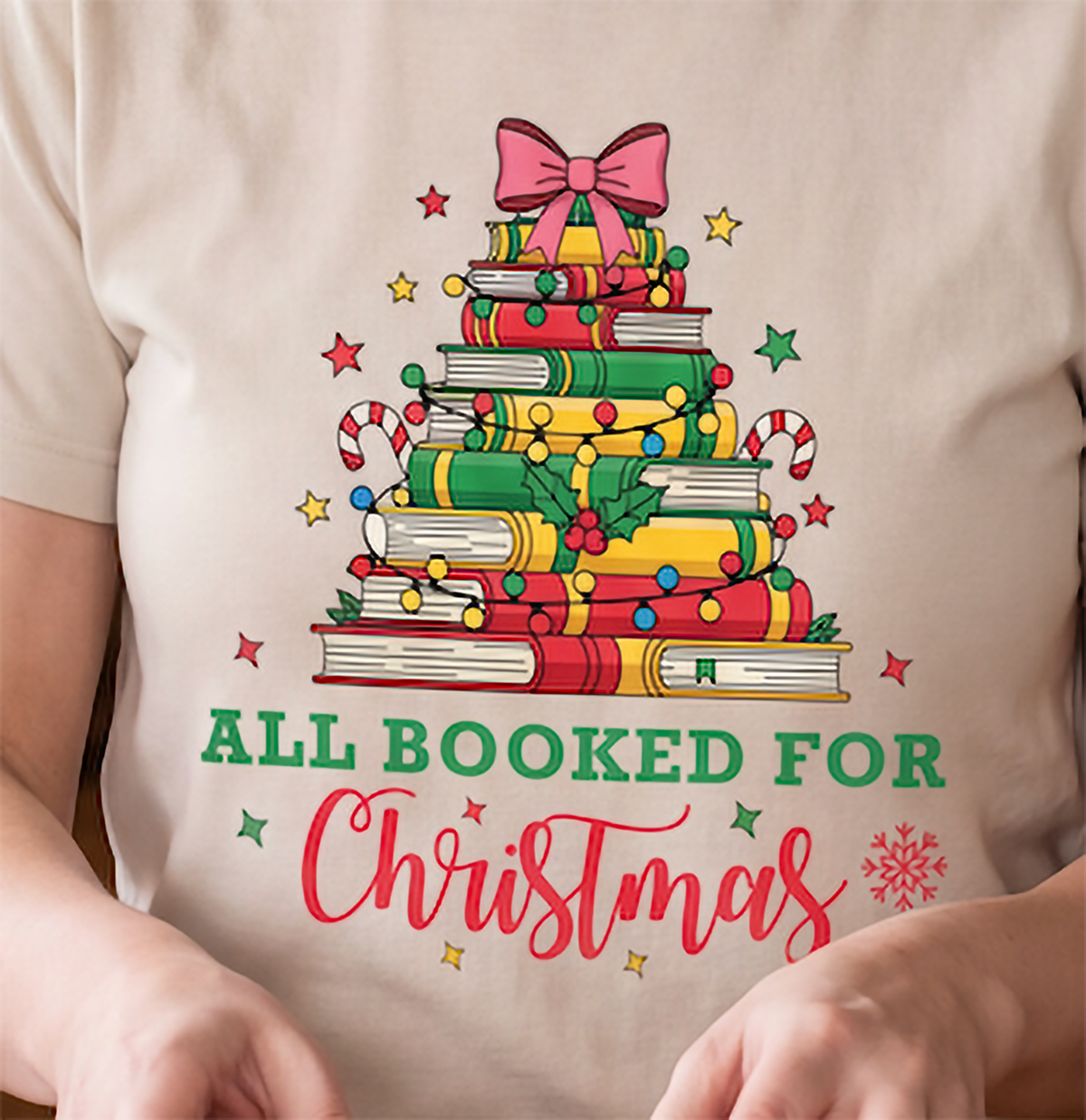 Close-up of sand All Booked for Christmas shirt showing colorful books, Christmas lights, candy canes and a bow.