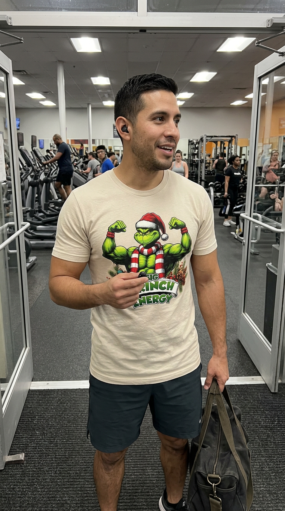Man wearing sand-colored Big Grinch Energy Christmas t-shirt at the gym featuring a muscular green Grinch holiday graphic.