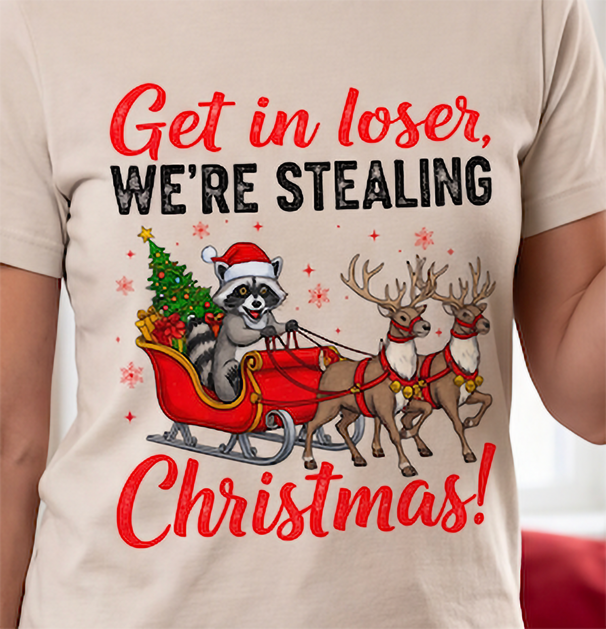 Close-up of Get in Loser We’re Stealing Christmas holiday raccoon design on a sand T-shirt