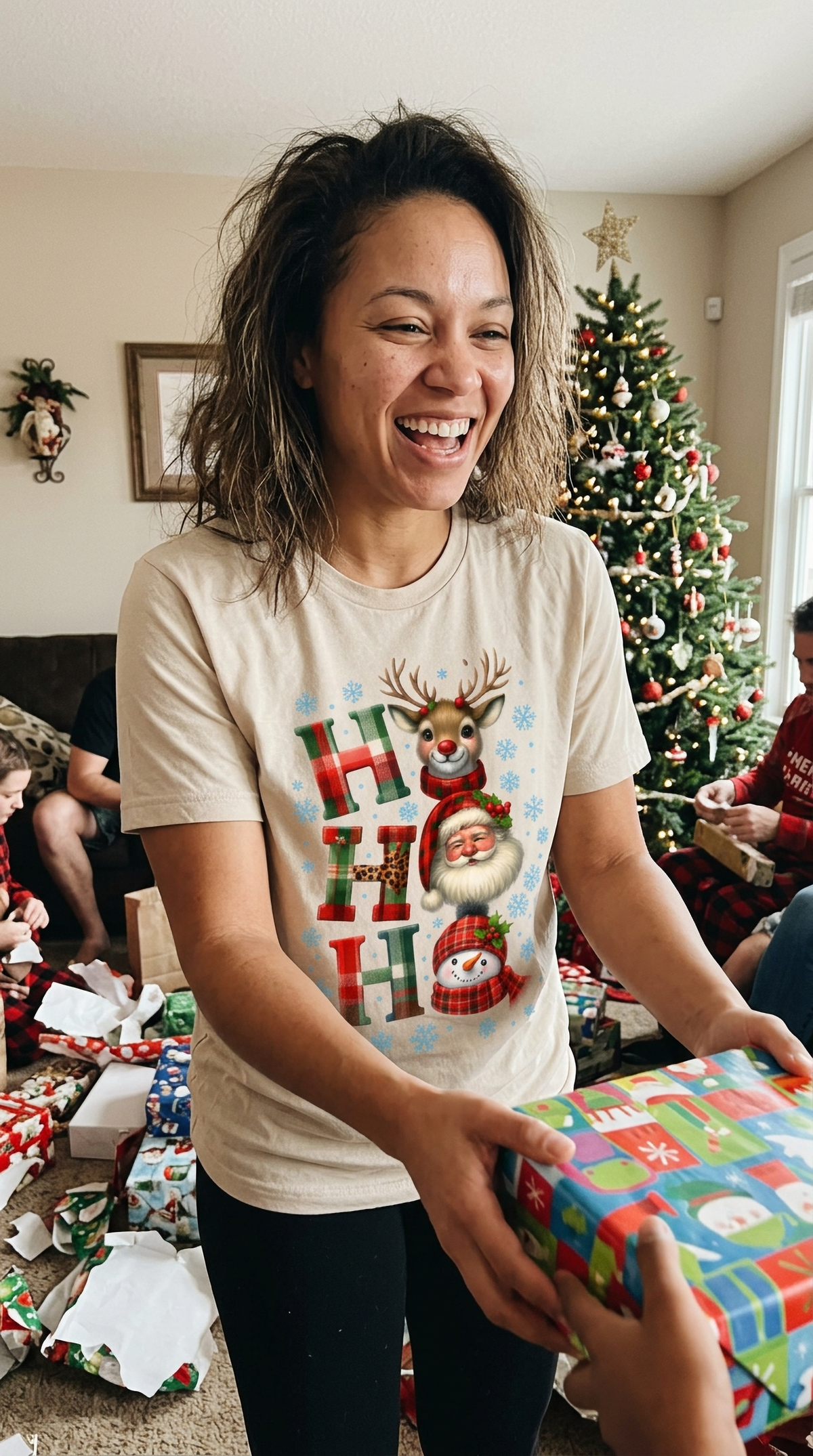 Smiling woman wearing sand-colored Ho Ho Ho Christmas t-shirt while wrapping Christmas presents.