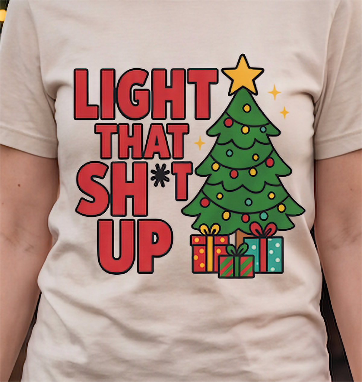 Close-up of sand Light That Shit Up Christmas shirt with bold lettering and festive tree graphic.