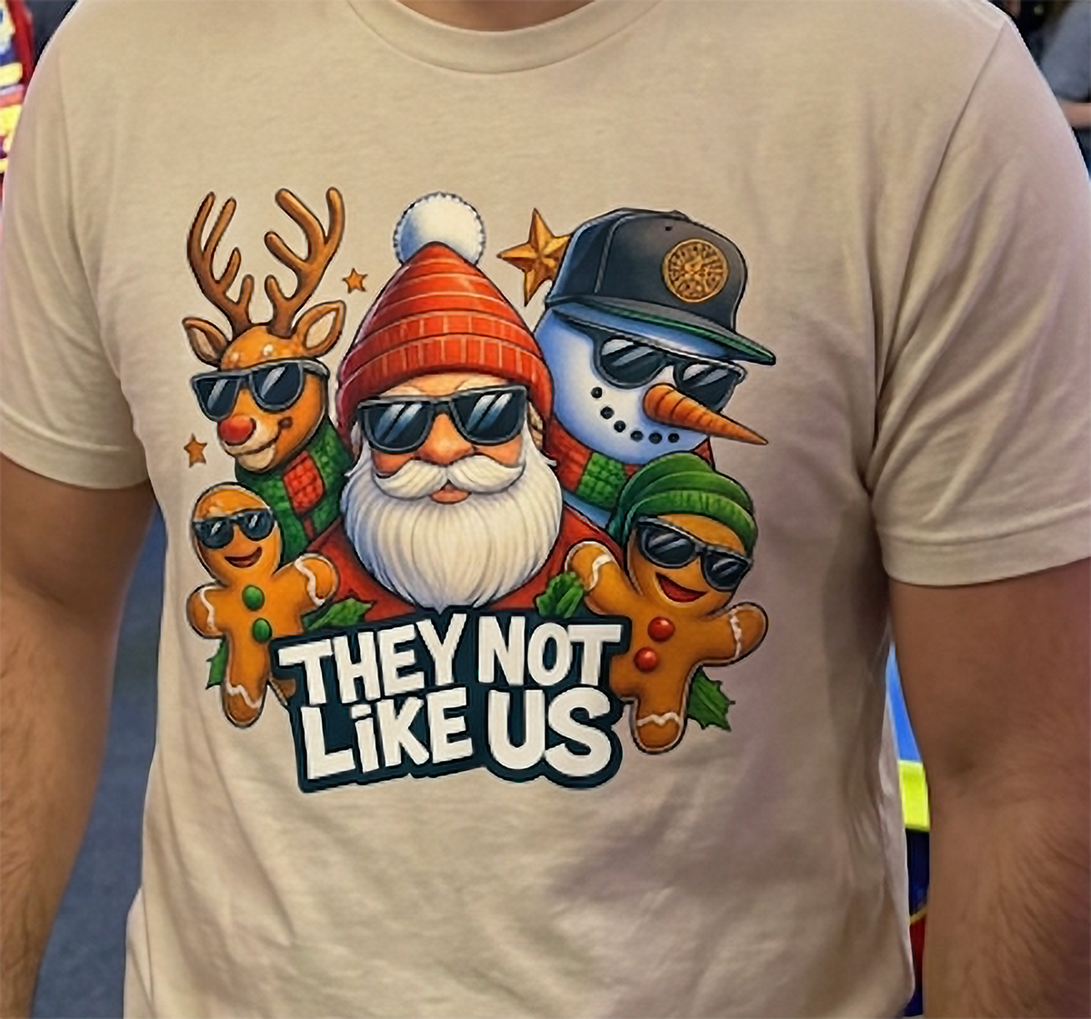 Close-up of sand Christmas t-shirt showing They Not Like Us graphic with playful holiday characters and bold lettering.