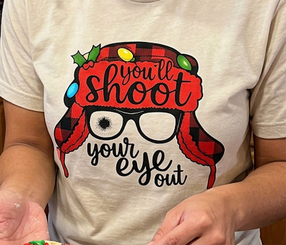 Close-up of sand Christmas graphic shirt displaying You'll Shoot Your Eye Out lettering and red winter hat design