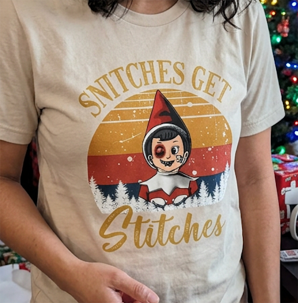 Close-up of Snitches Get Stitches Christmas elf design printed on a sand-colored unisex graphic tee.
